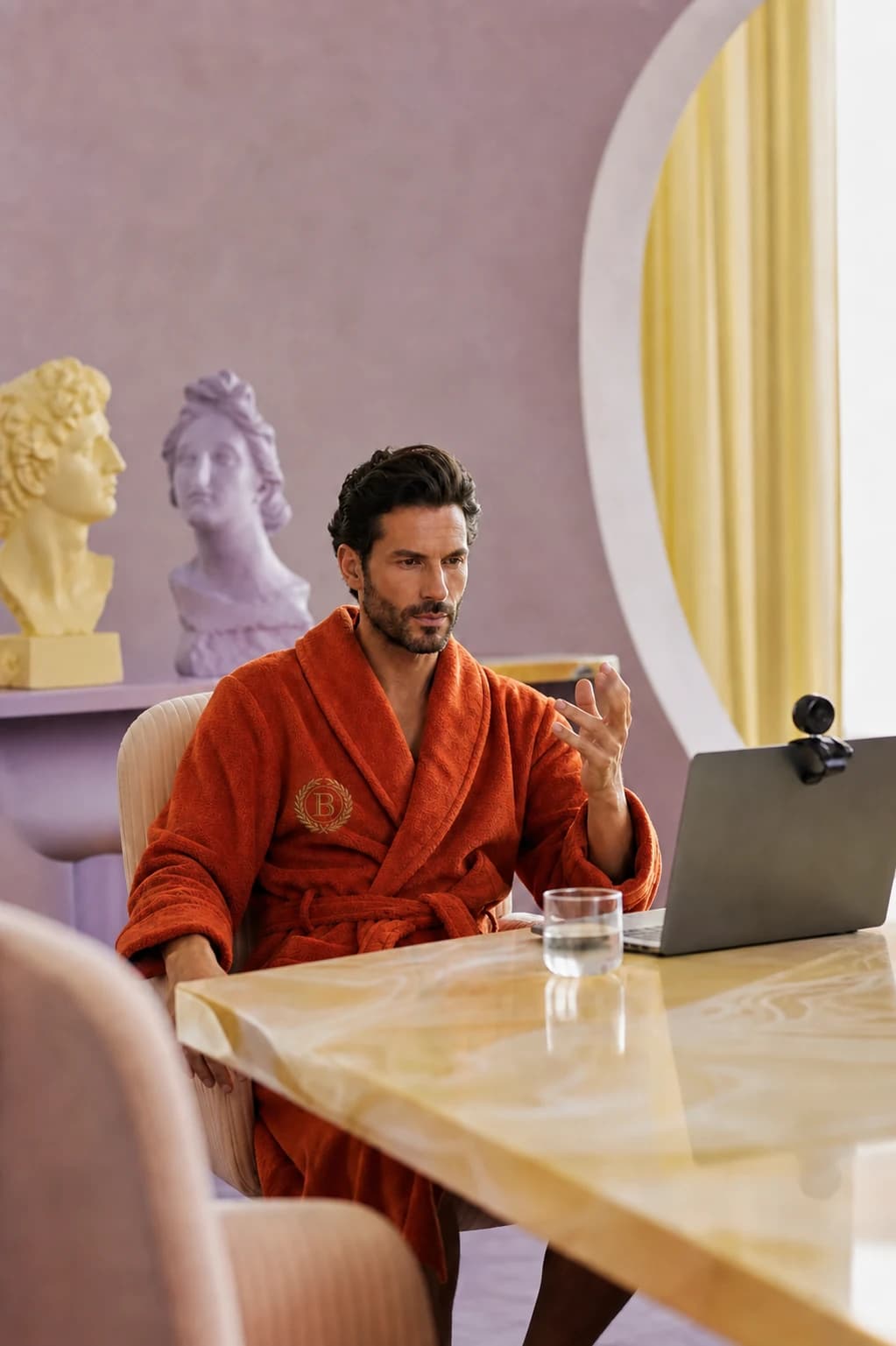 A model in a burnt-orange robe attends an absurd luxury board meeting.